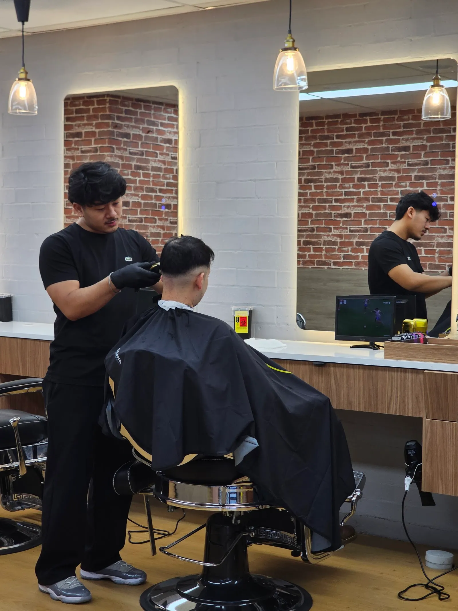 A barber cutting a client’s hair in front of lit mirrors and a brick wall